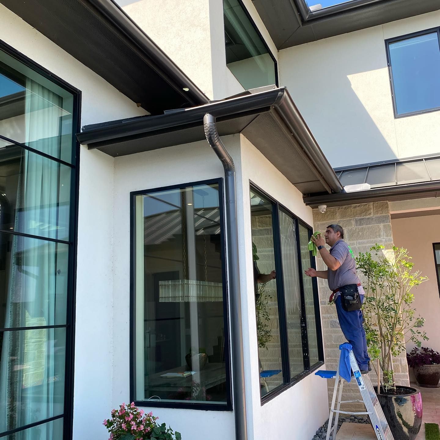 Man cleaning windows on a white house with black trim. He is on a ladder and wearing a blue shirt.