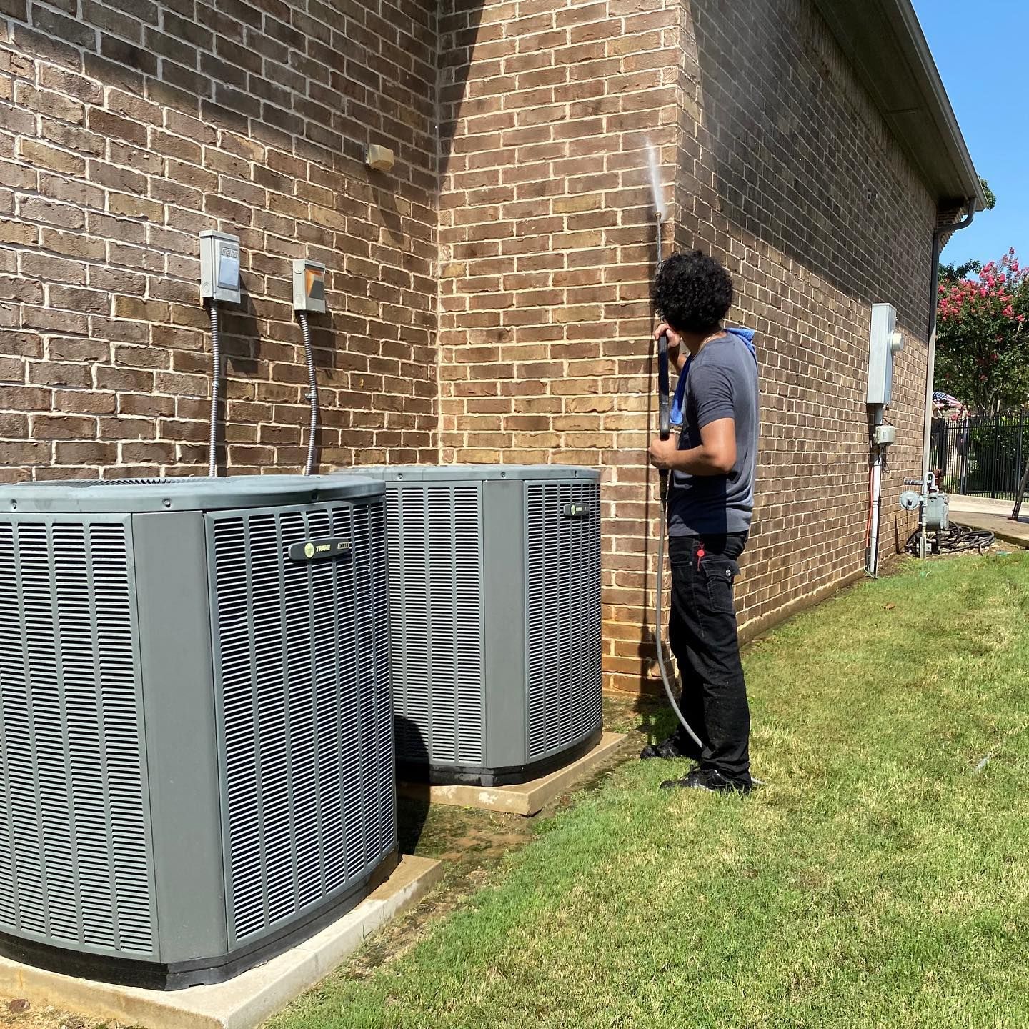 Man power washing a brick wall next to two AC units on a sunny day.