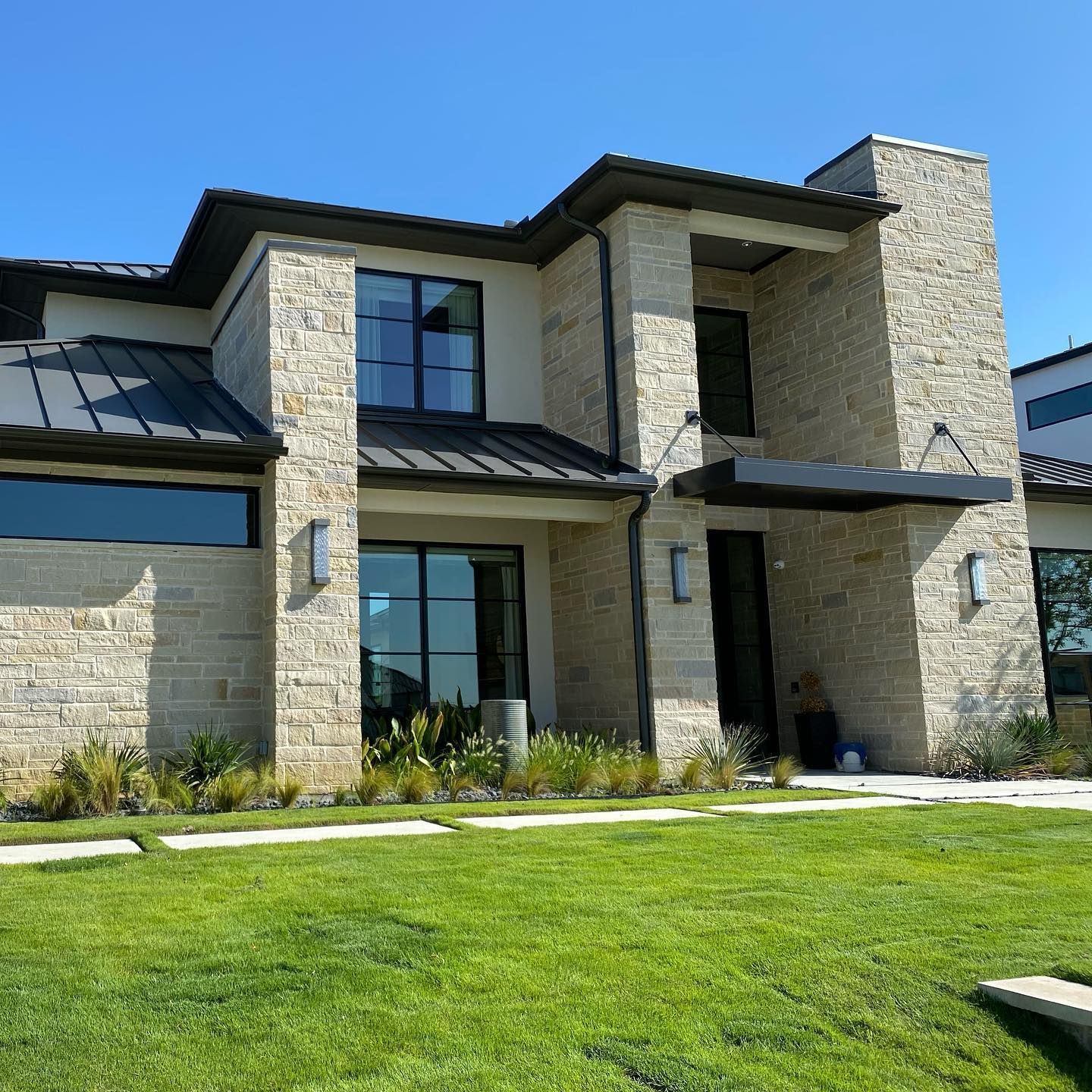 Modern two-story house with light stone walls, black trim, and a green lawn under a clear blue sky.