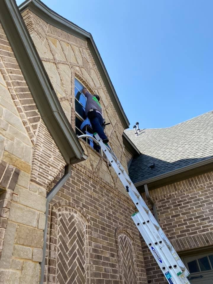 Person on ladder cleaning a window on a brick house with a dark roof, against a bright blue sky.