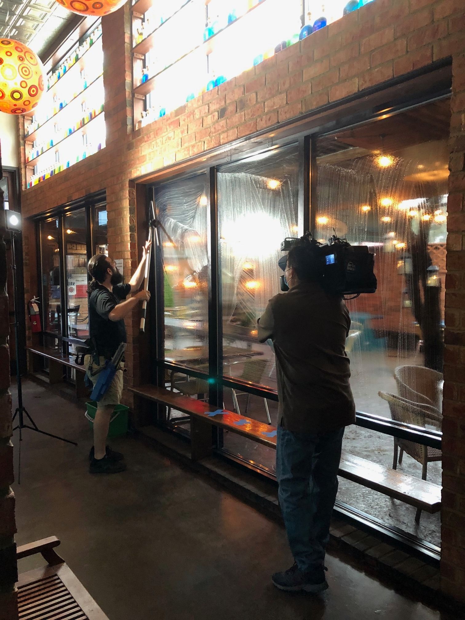 Two people film through a window. One cleans the glass; the other operates a camera. Inside a brick building.