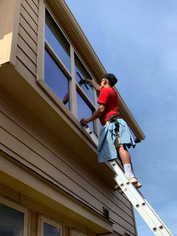 Person washes window on ladder, red shirt, blue sky, beige siding.