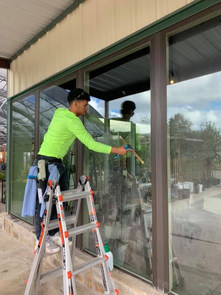 Person in neon green shirt cleans a large window with a squeegee while standing on a ladder.