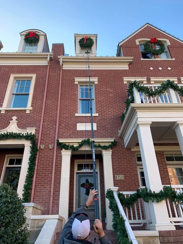 Person adjusting a pole to hang holiday decorations on a brick building with wreaths and garland.