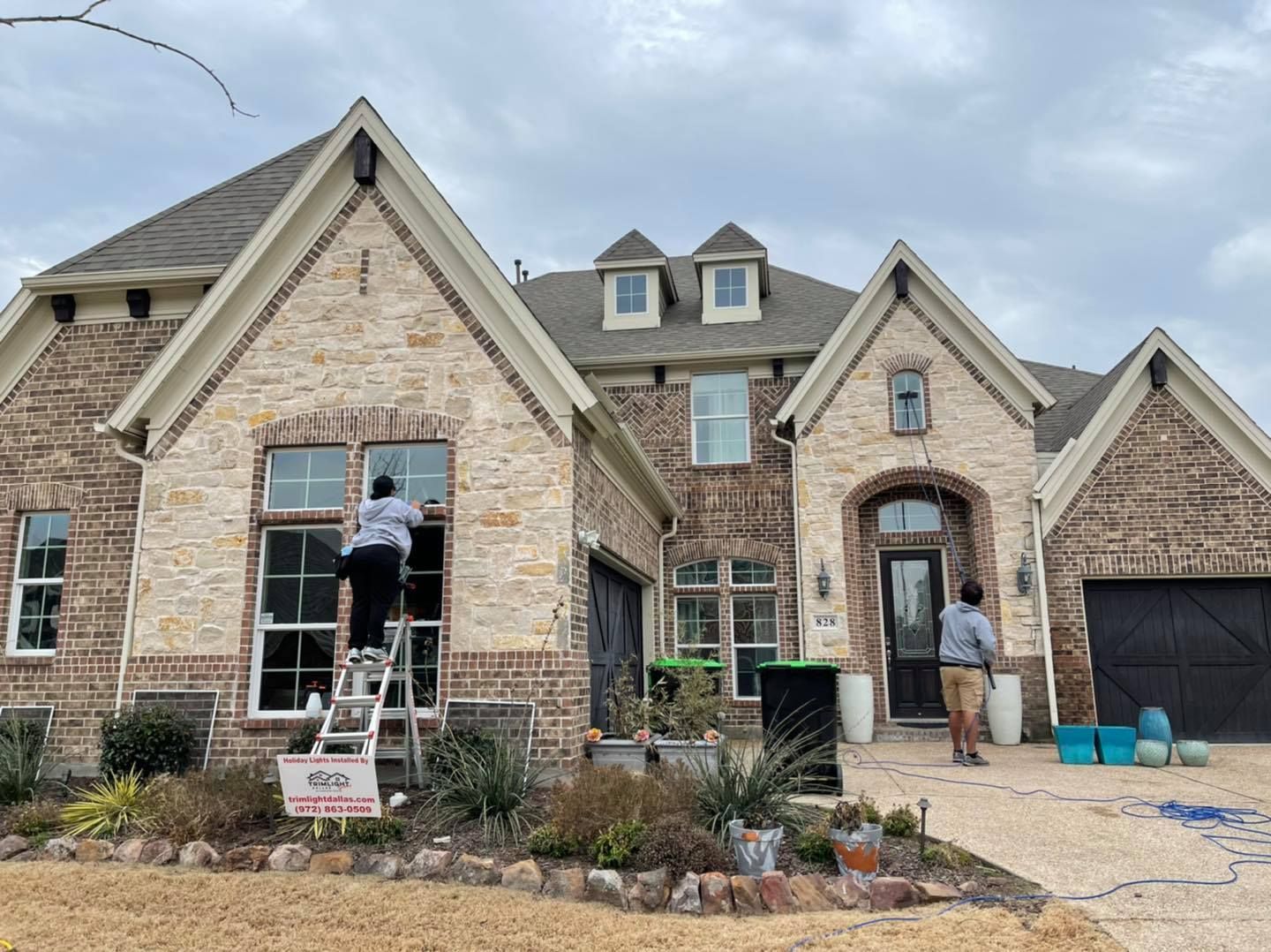 Two people cleaning windows of a large brick house on an overcast day. One on a ladder.