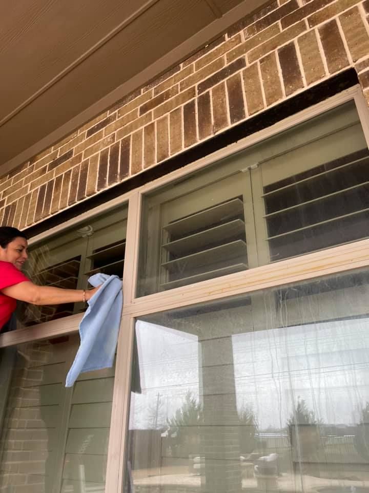Woman cleaning a window outdoors with a blue cloth. She wears a red shirt, and a brick wall is above.