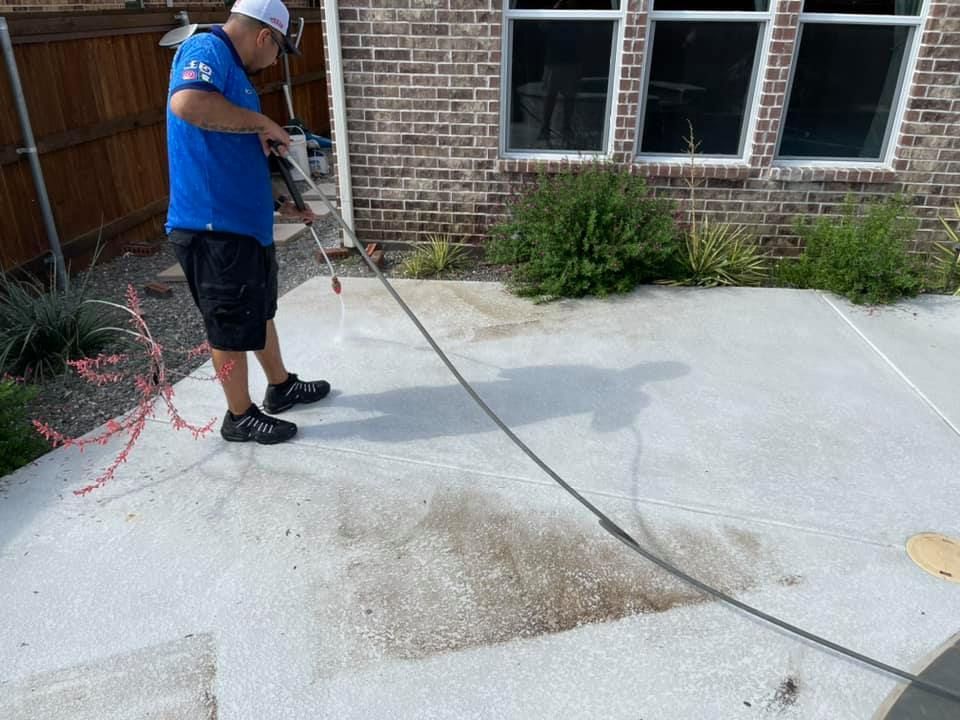 Man in blue shirt pressure washing a concrete patio next to a brick house.