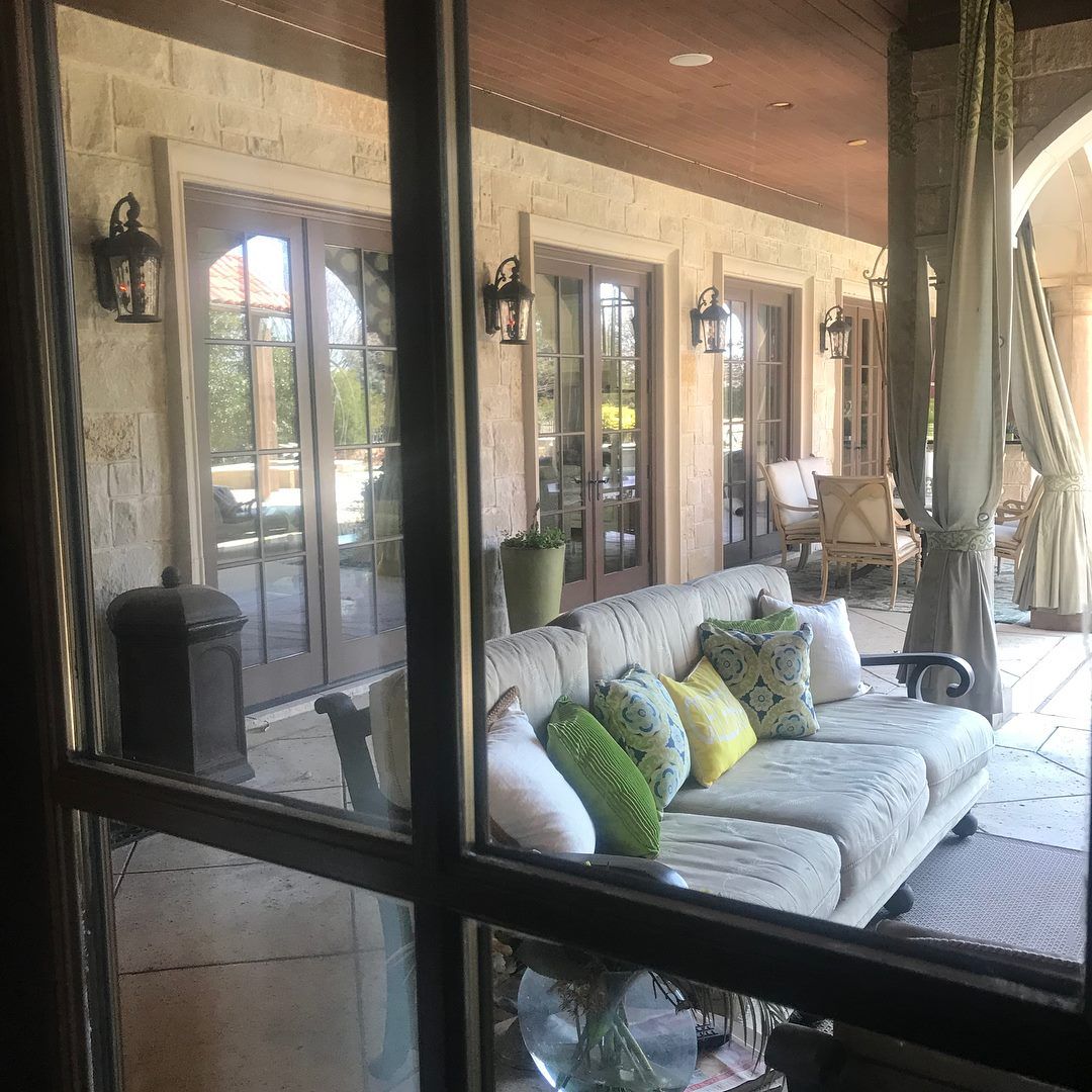 A covered patio with a beige couch, colorful pillows, and stone walls, viewed through a window.