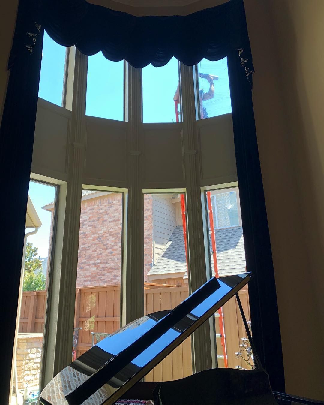 Black grand piano beneath a large window with blue sky view, dark curtains and valance.