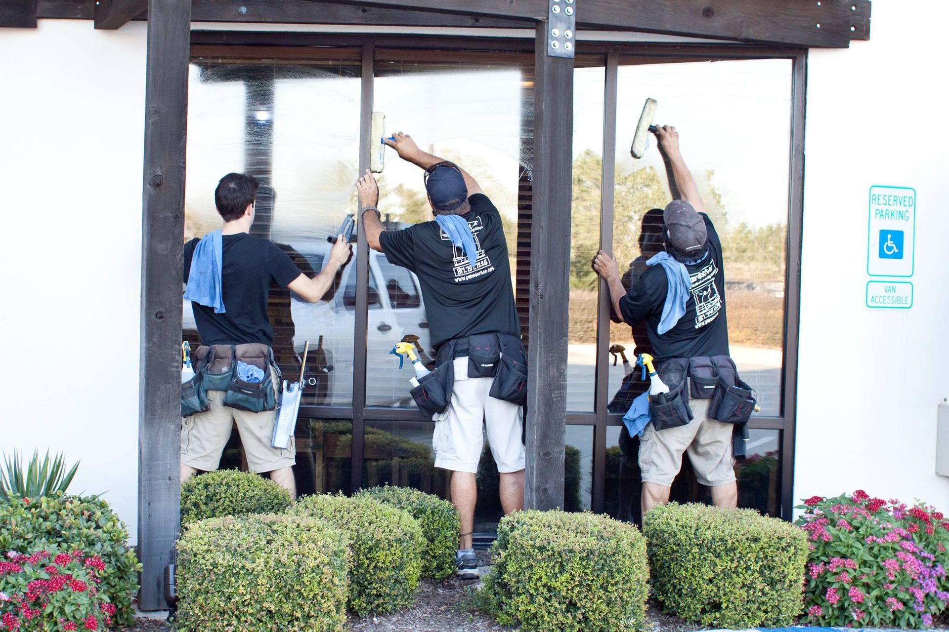 Three window cleaners squeegee an exterior window; they wear dark shirts, shorts, and tool belts.