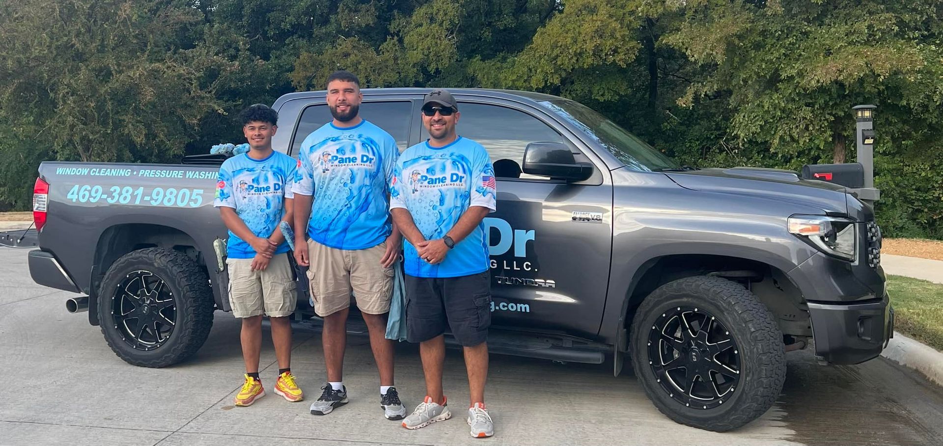 Three men stand beside a gray pickup truck. They wear matching blue shirts with a water design.