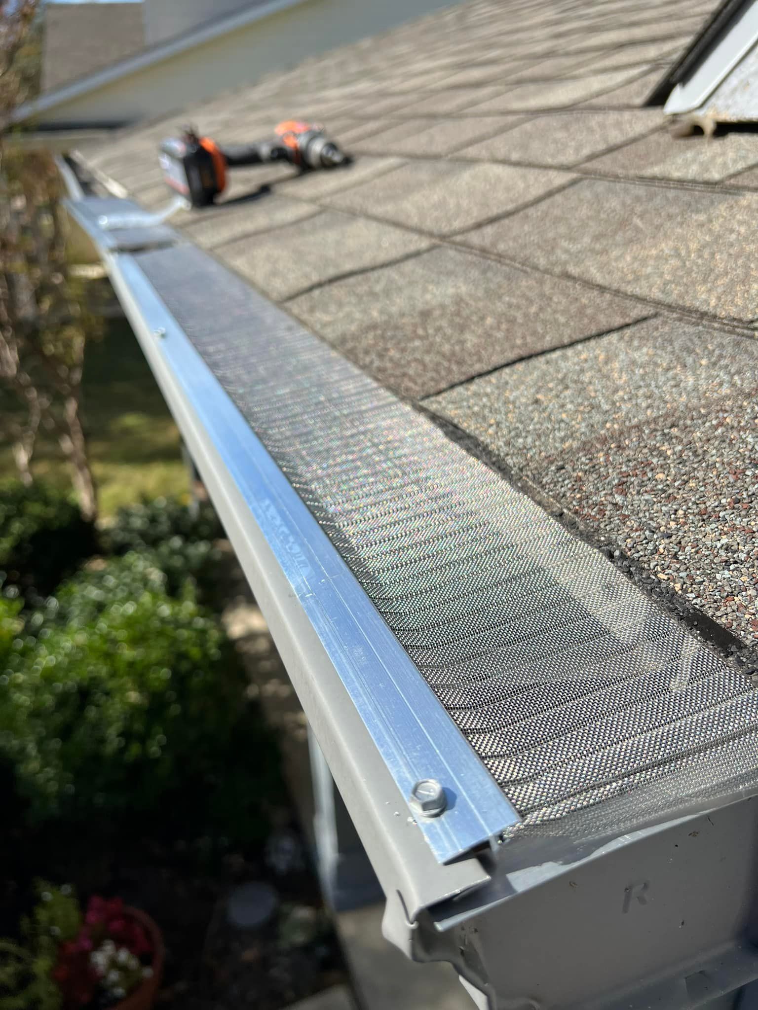 Gutter with installed mesh screen, secured with screws. A drill sits on roof above.