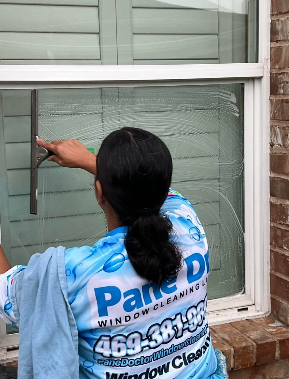 Woman cleaning a window with a squeegee; wearing a Pane Doctor window cleaning shirt outside a brick building.
