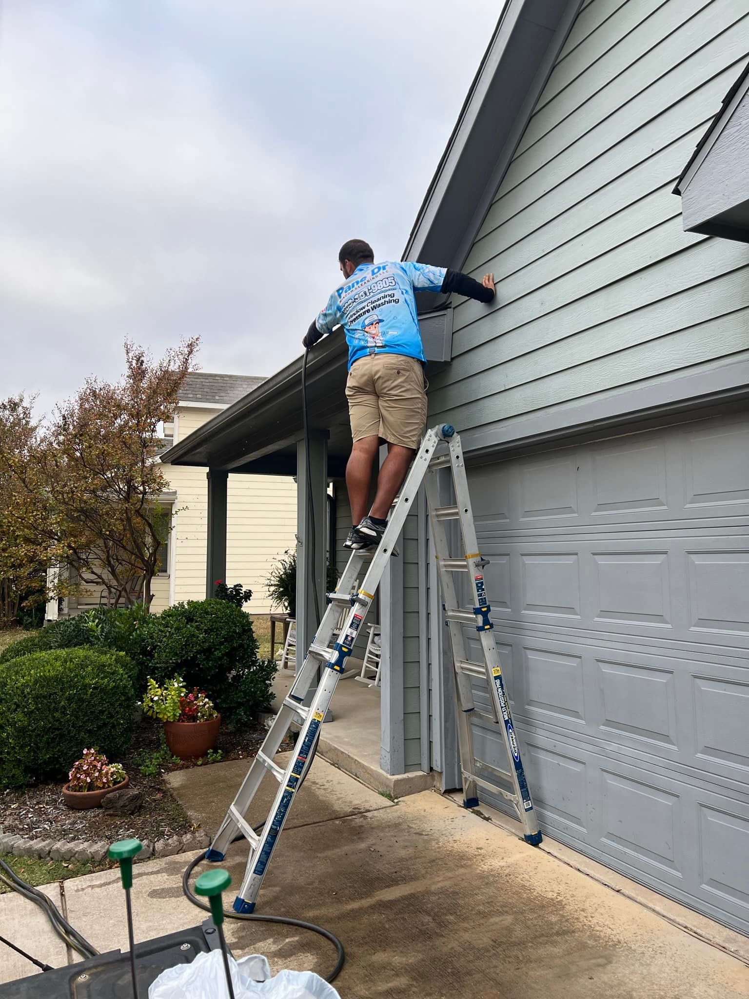 Man on a ladder near a gray house, working on the siding. The sky is overcast.