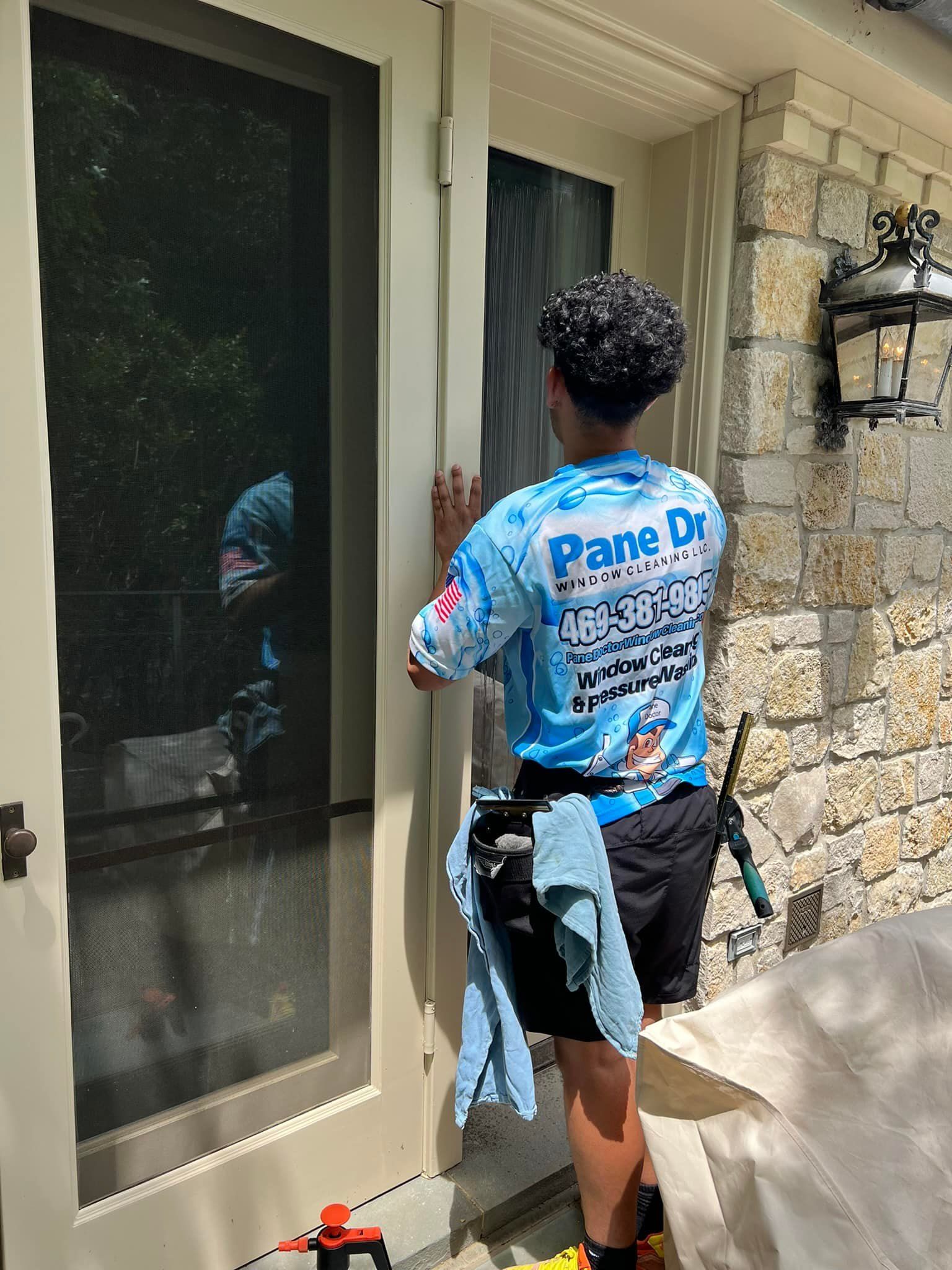 Window cleaner working on a door; light-blue shirt, black shorts, stone wall background.