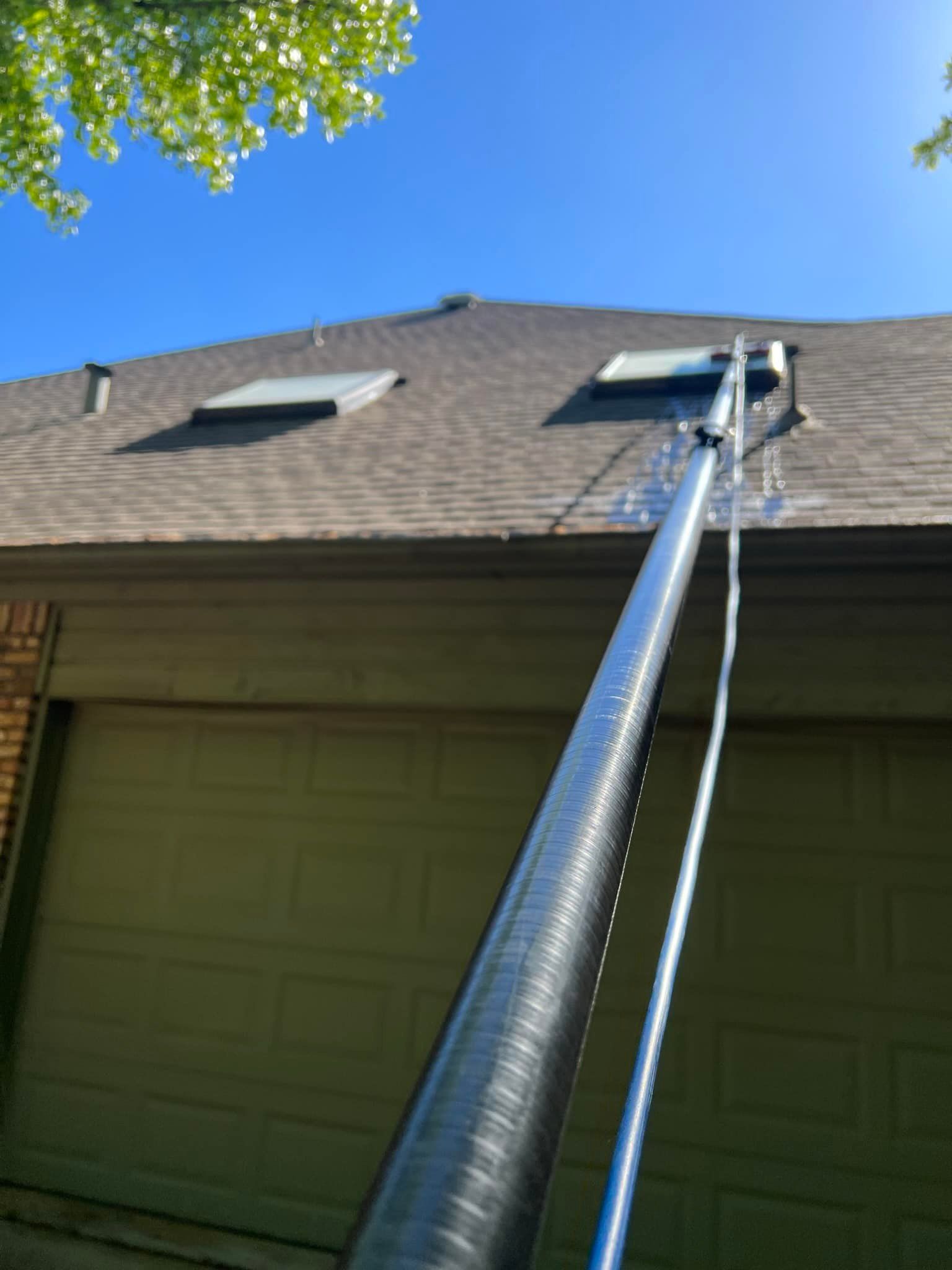 A long, silver gutter cleaning tool extends up to a brown shingled roof above a tan garage.