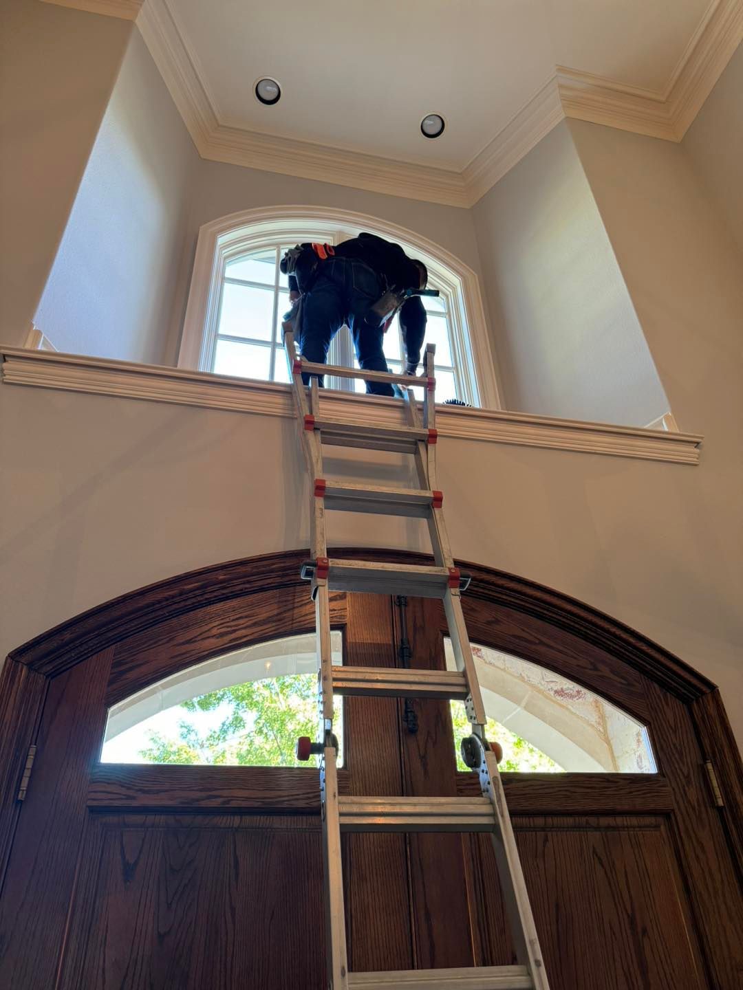 Person on ladder cleaning high window in a foyer with ornate molding and dark wooden door.