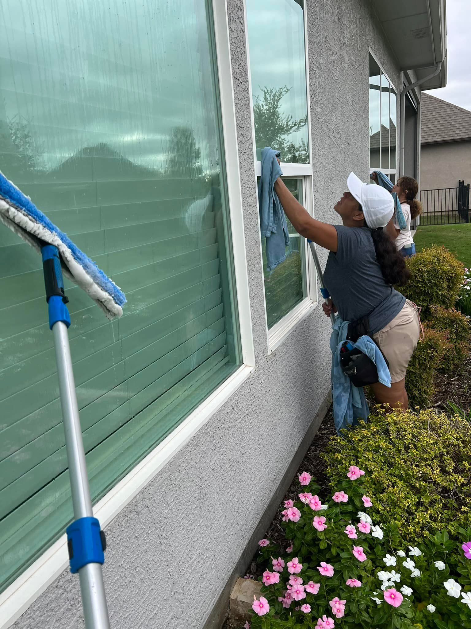 Woman in cap cleaning window, outside, with cleaning tools, flowers nearby.