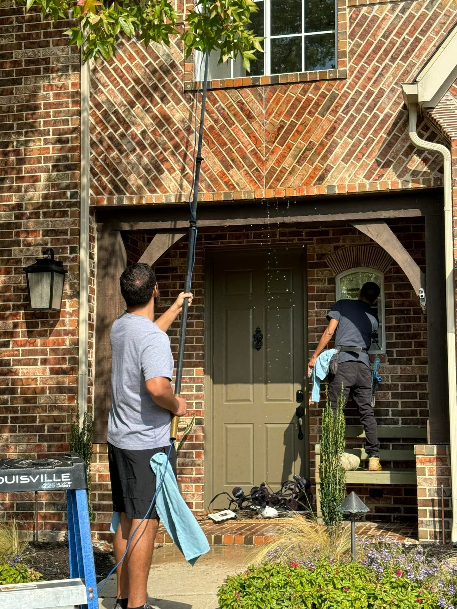 Two people washing windows of a brick house. One uses a pole, the other cleans by hand.