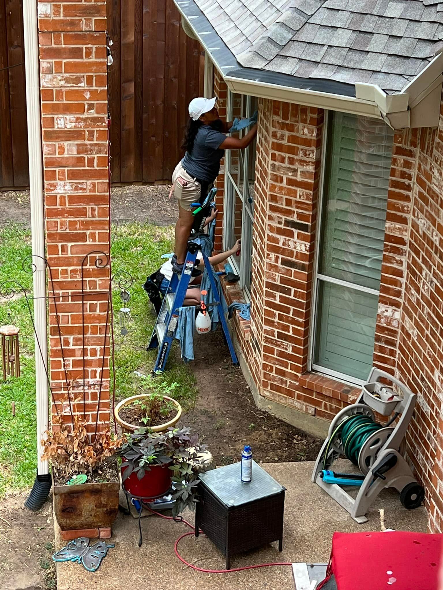 Person cleaning windows on a ladder next to a brick house, using a spray bottle and cleaning tools.