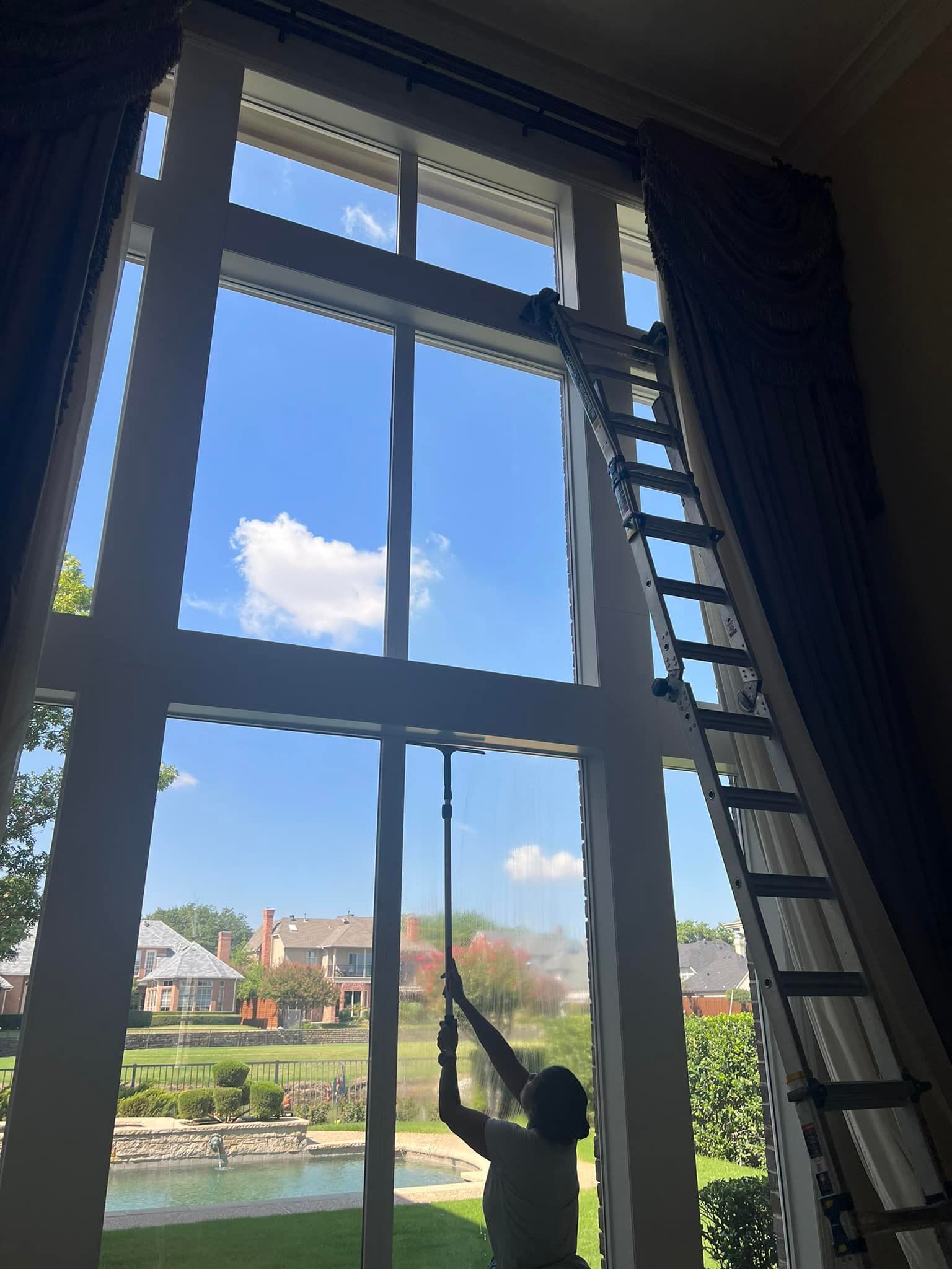 Person on ladder cleaning tall window, blue sky and houses visible.