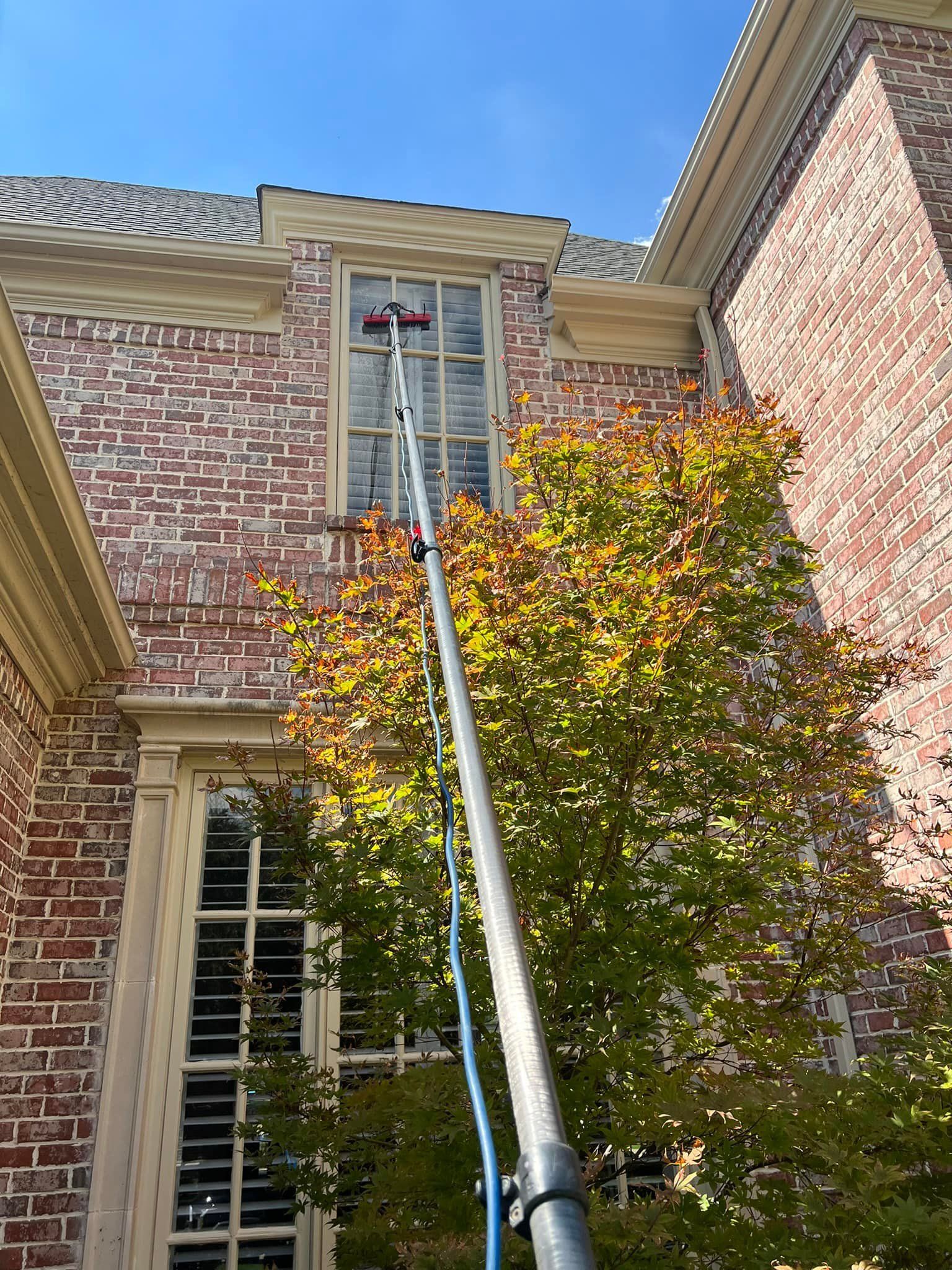 A person cleaning a window on a brick building with a long-handled brush; fall foliage.