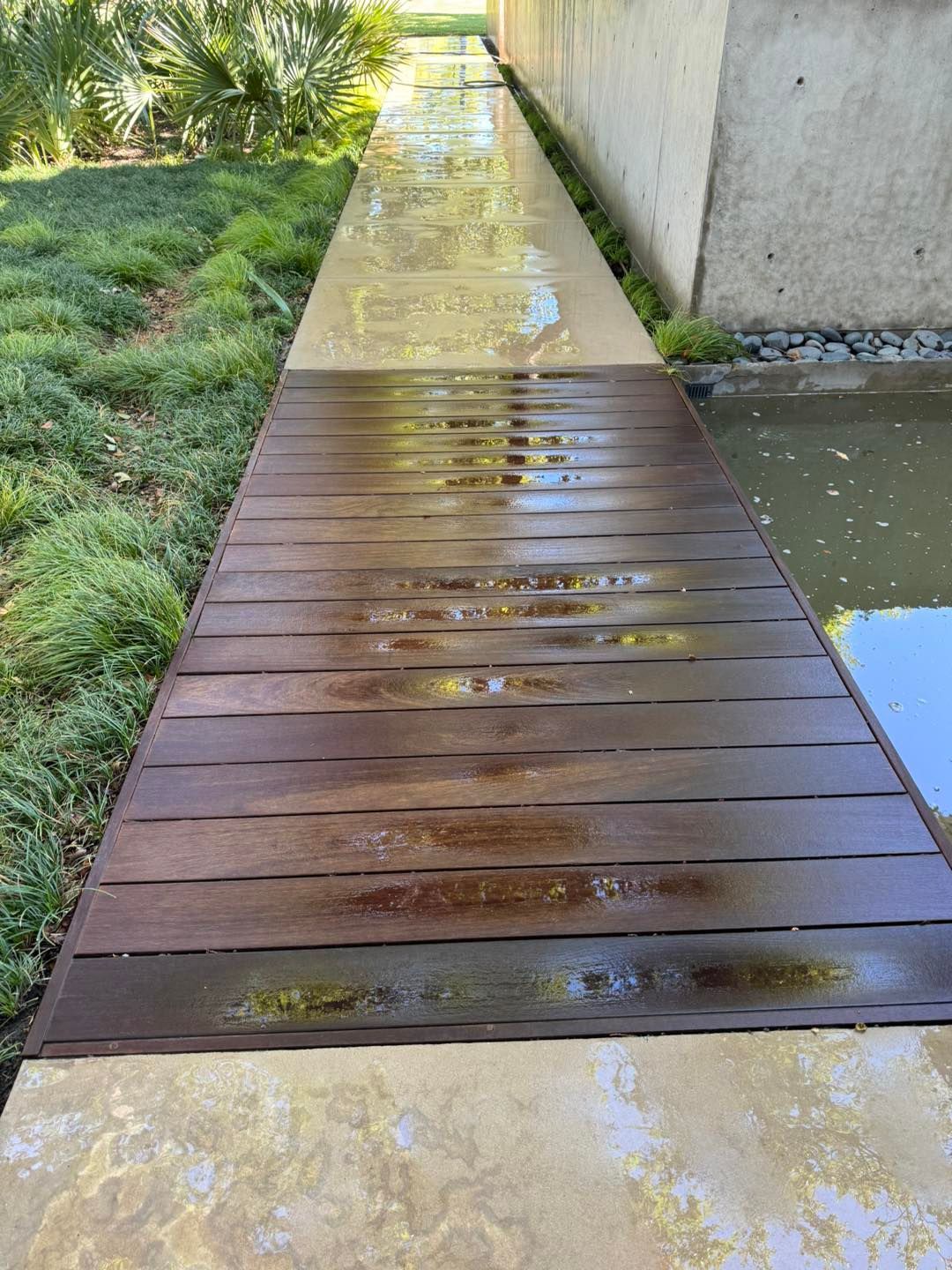 Wooden walkway bridge over water, beside grass and concrete.