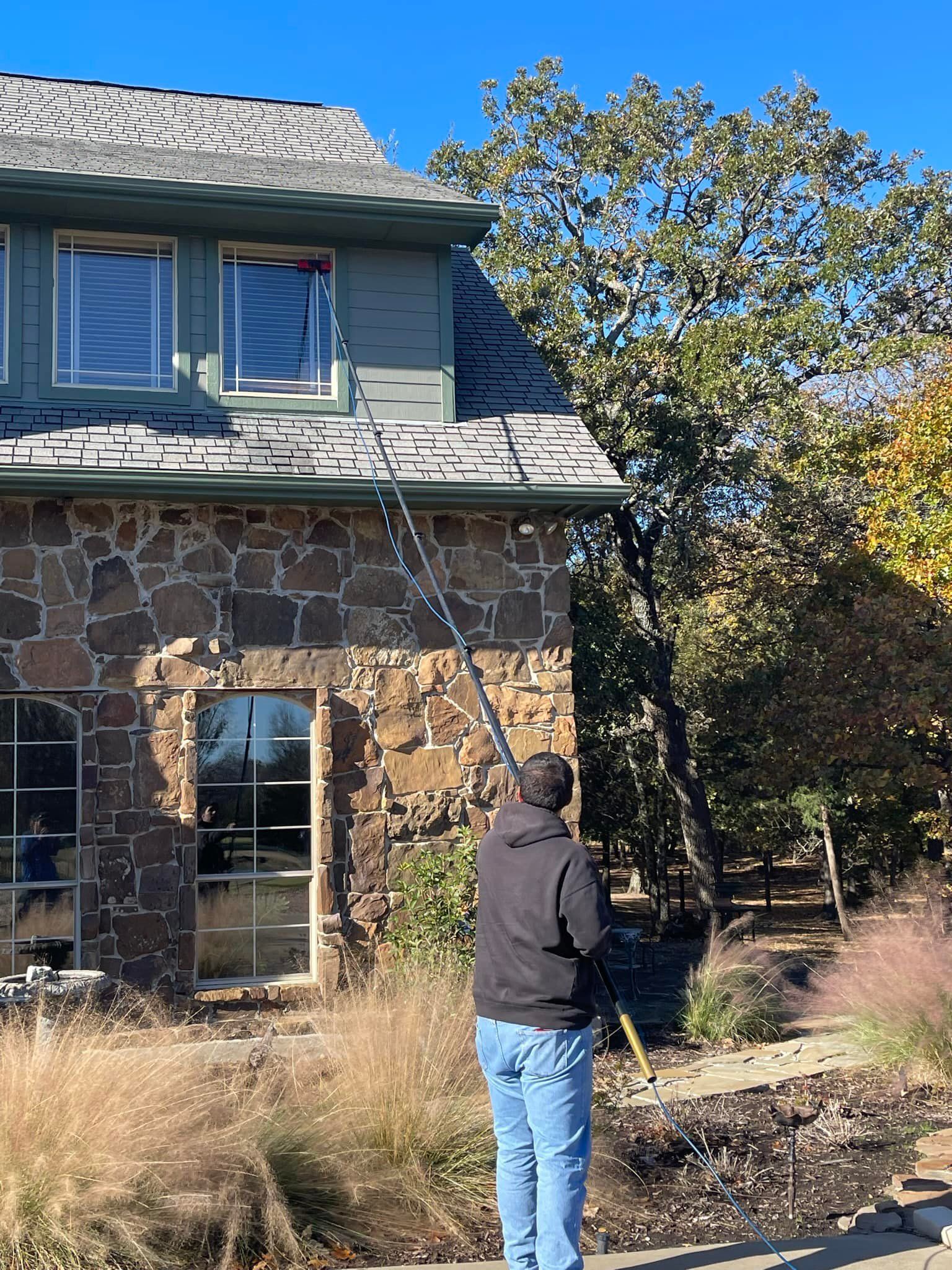 Person cleans gutters on a house with stone and green siding, using a long-handled tool. Clear blue sky.