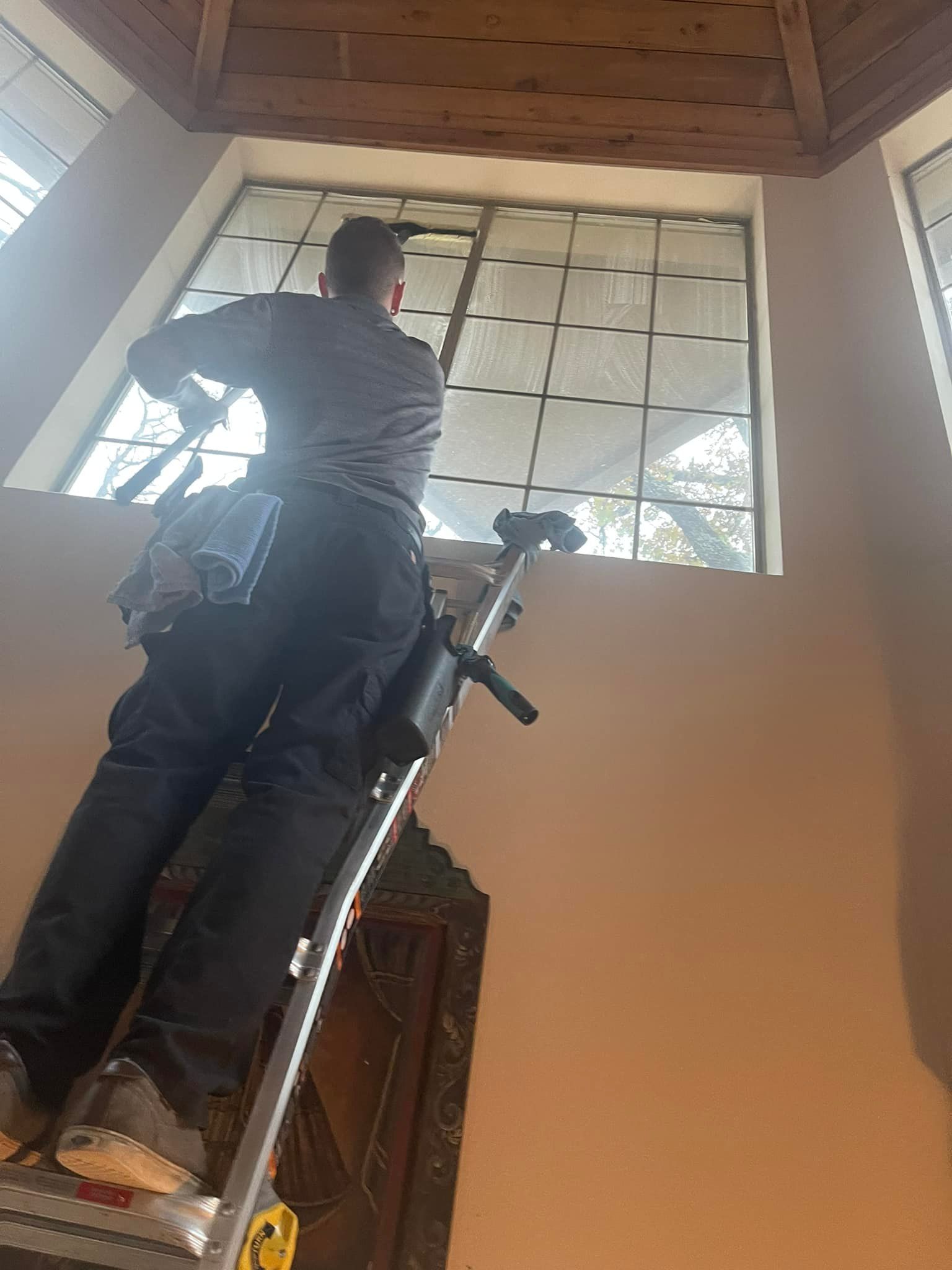 Man on ladder cleaning a large, multi-pane window indoors.