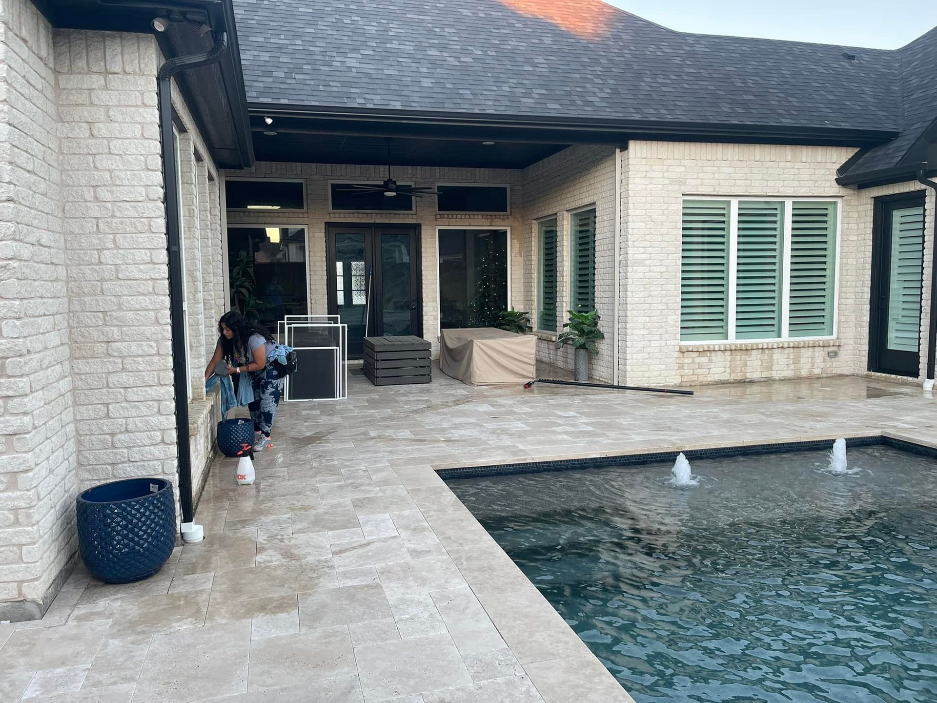Person near a pool on a patio of a brick home. Fountain sprays water. Evening sunlight.