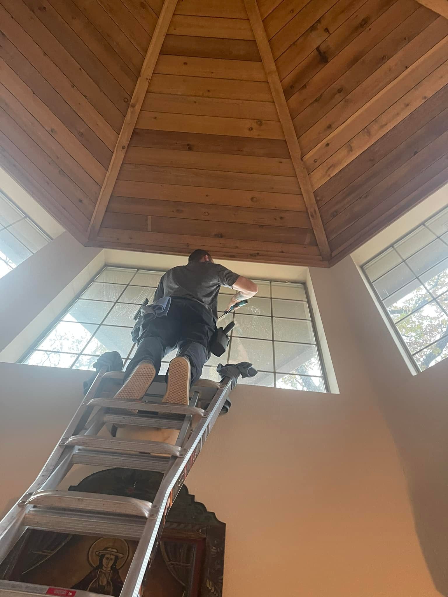 A person on a ladder cleaning a high window inside a building with a wood ceiling.