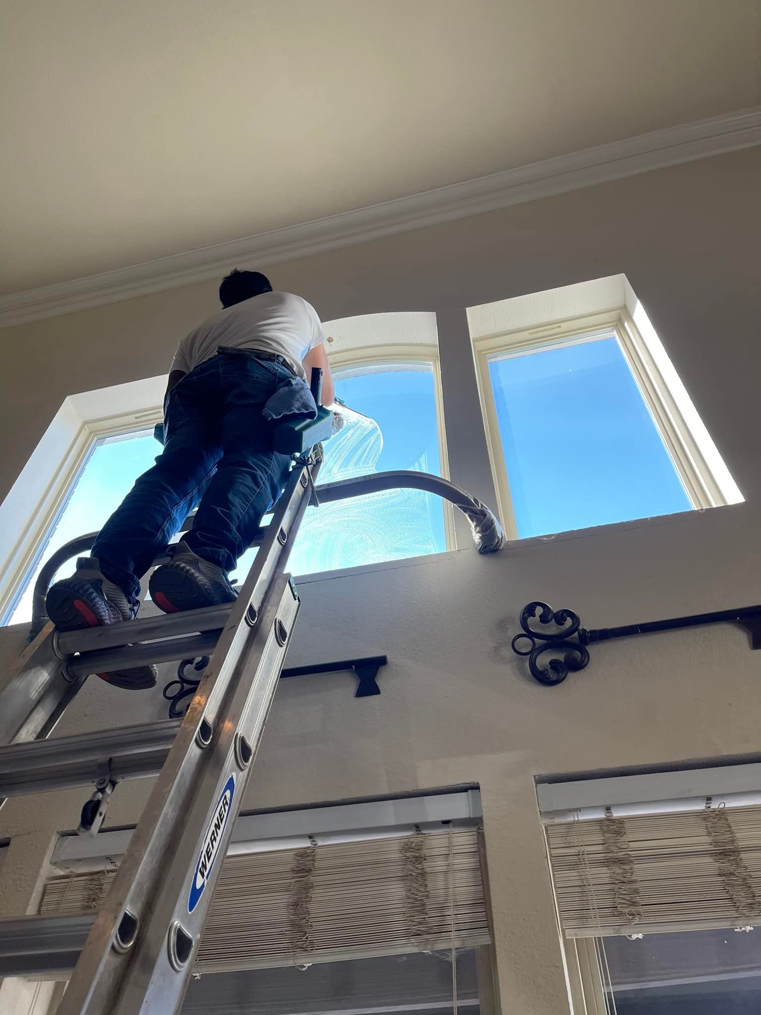 Man on ladder cleaning a tall window, blue sky visible.