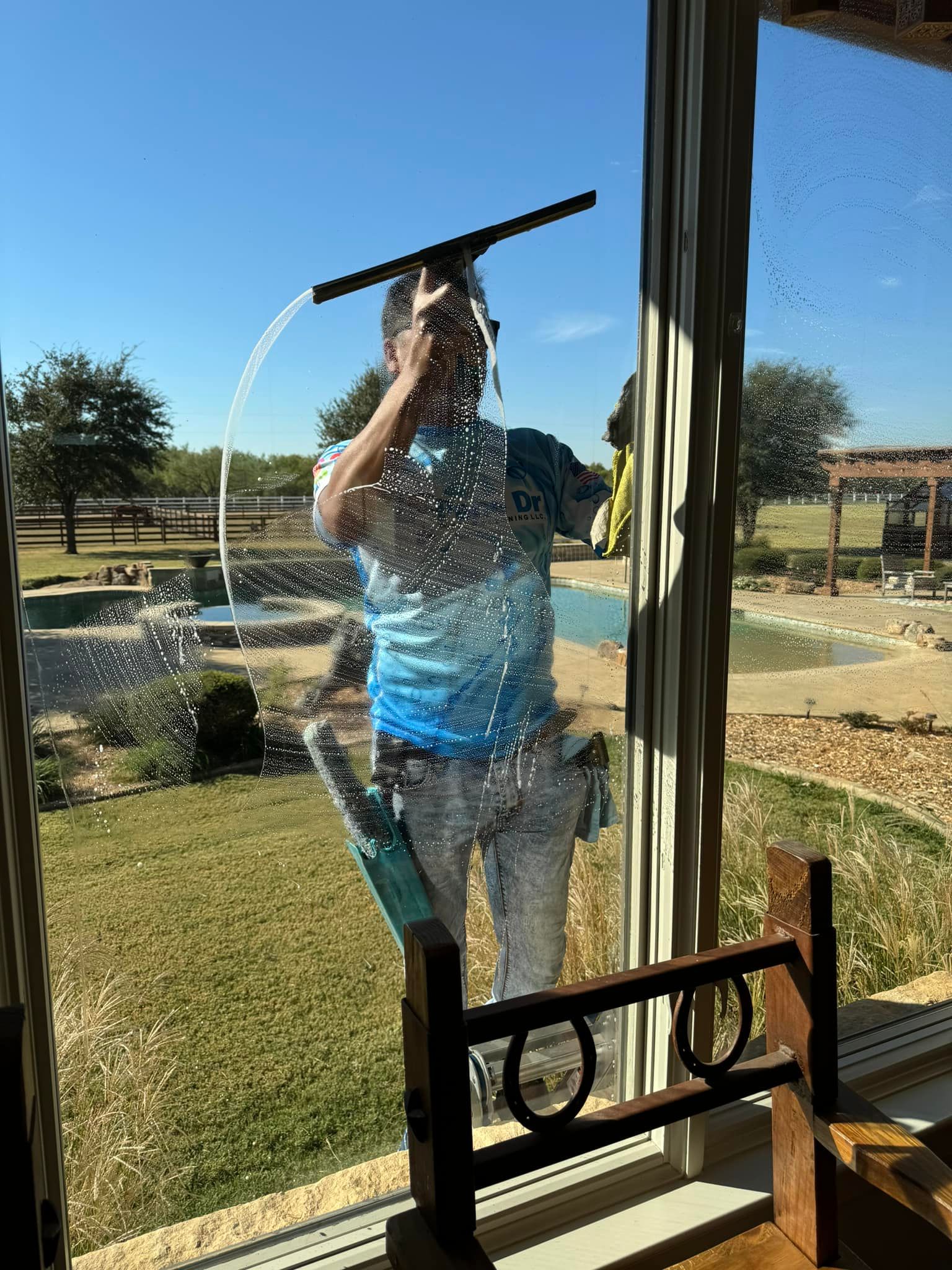 Person cleaning a window with a squeegee. Blue sky, green lawn, and a pool in the background.