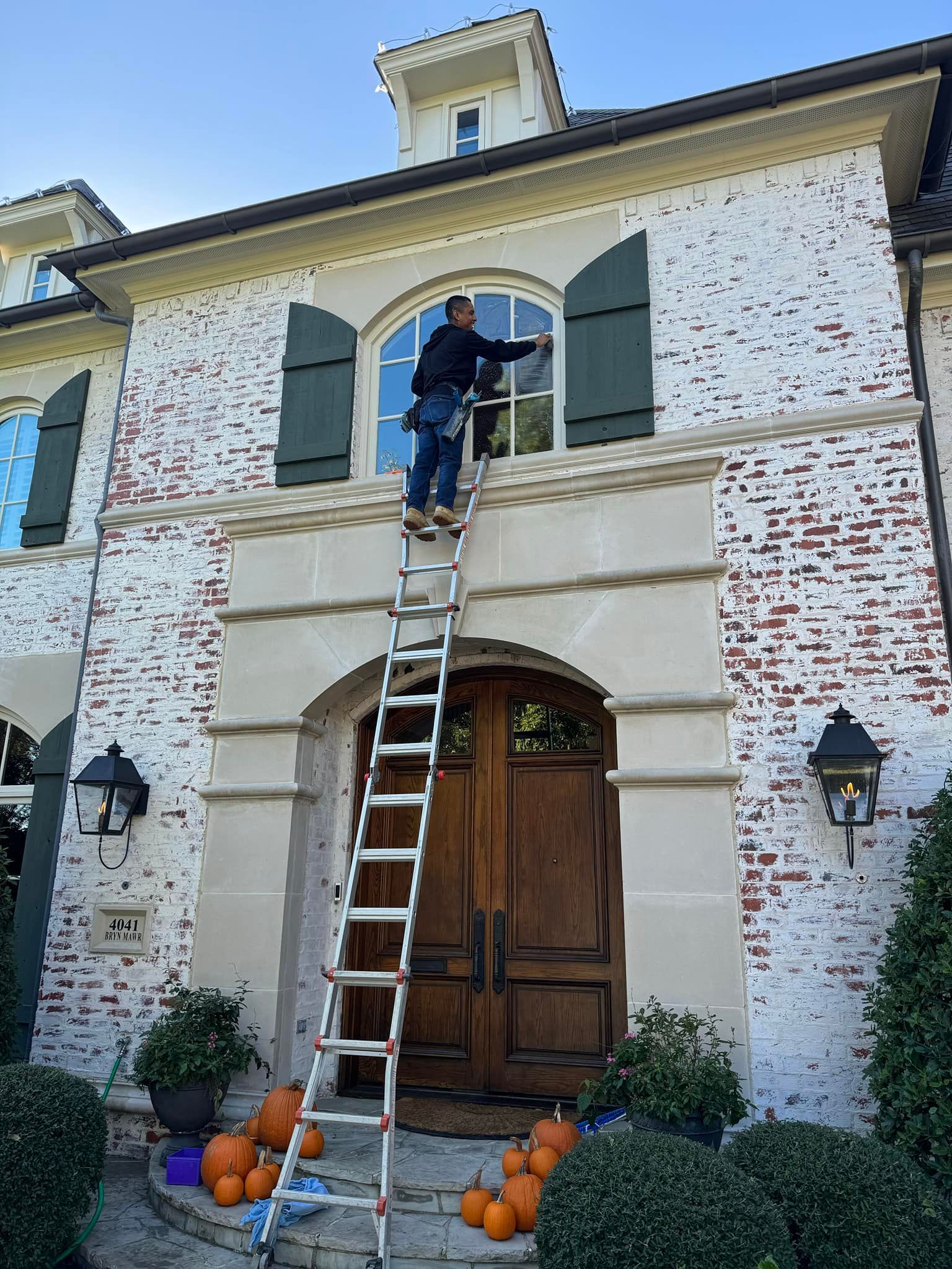 Person on a ladder washing a window on a brick house with green shutters; pumpkins on the porch.