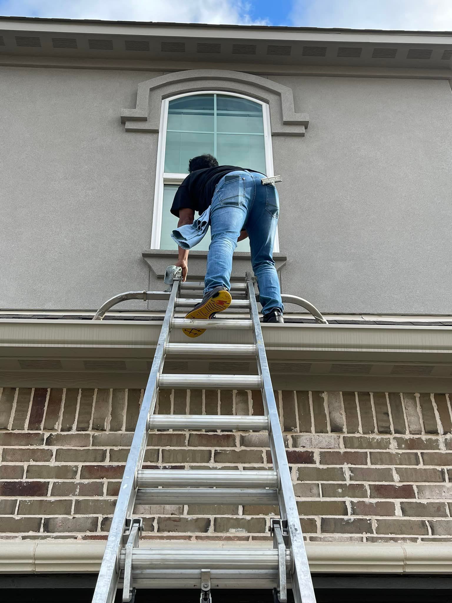 Man on a ladder, working on the exterior of a brick building, near a window.