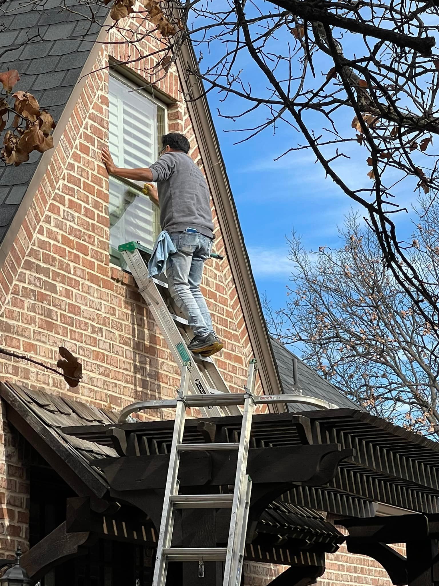 Person on a ladder cleaning a window on a brick building with a blue sky in the background.