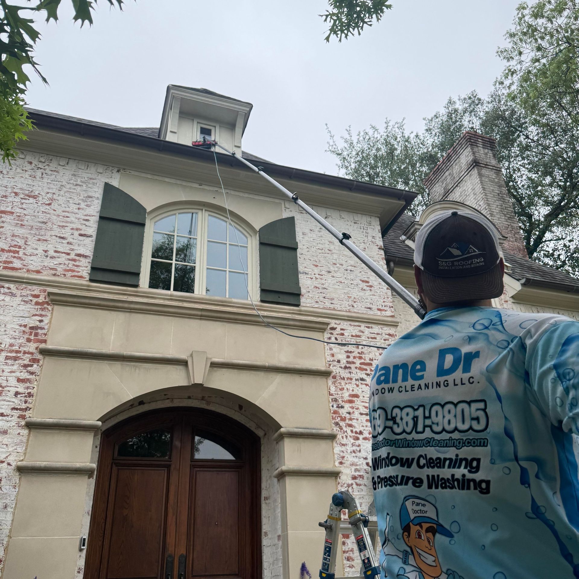 Man cleaning a window on a two-story brick house with a long pole, green shutters, and a cloudy sky.