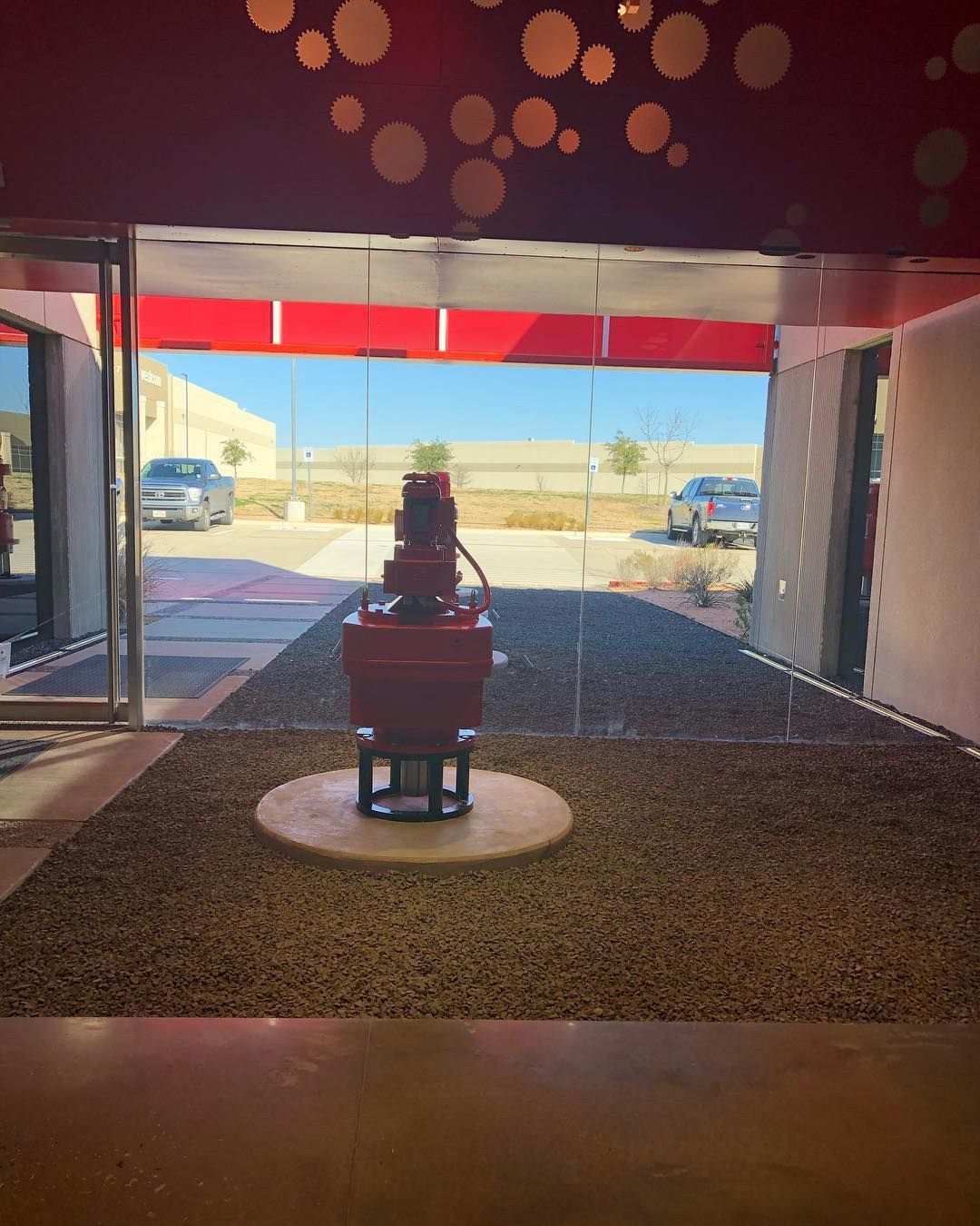 A red machine on a circular base, seen through a window. Gravel ground, cars in the background.