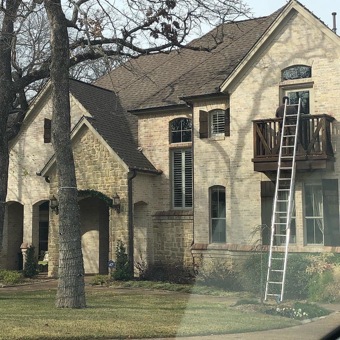 House with a ladder leaning against the balcony, beige brick, and brown roof in front of a tree.
