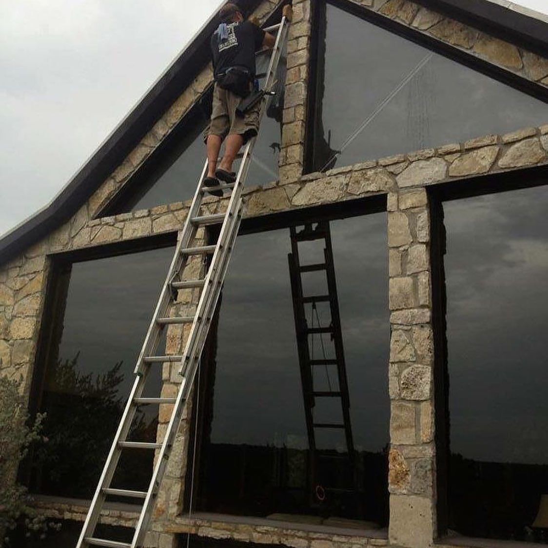 Man on ladder cleaning large, dark windows of a stone house with an A-frame roof.