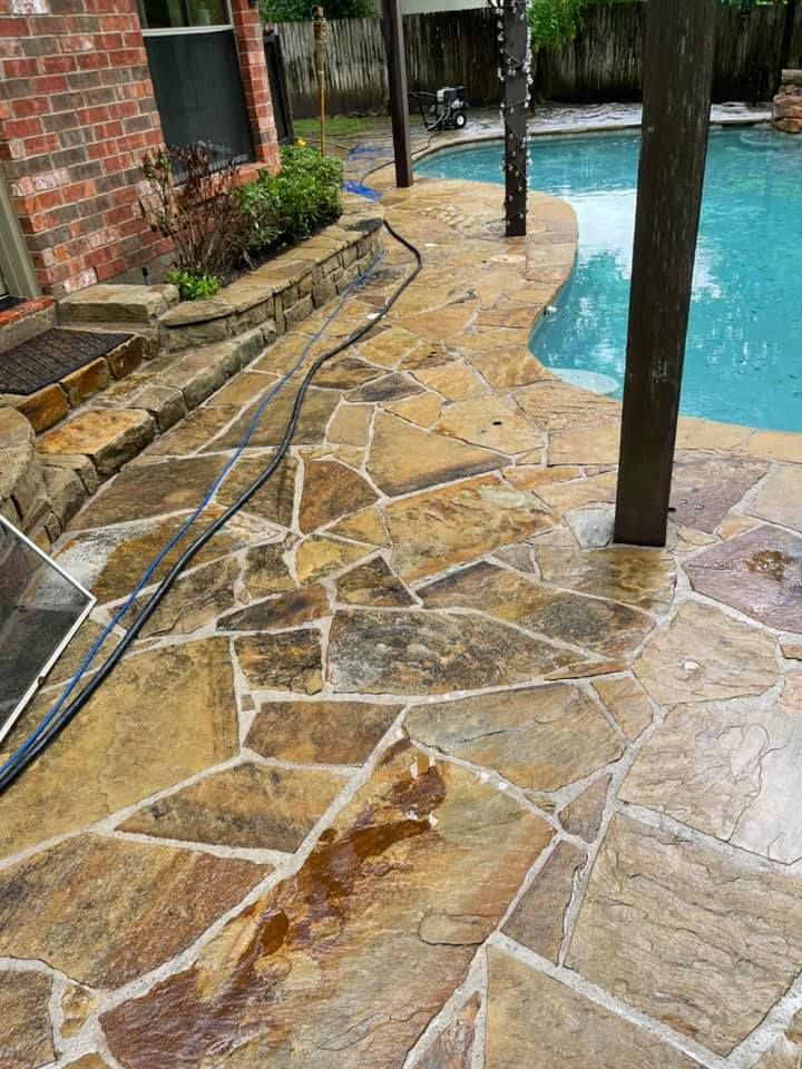 Stone patio around a pool with a brick house. The patio is wet, with brown and tan stones.
