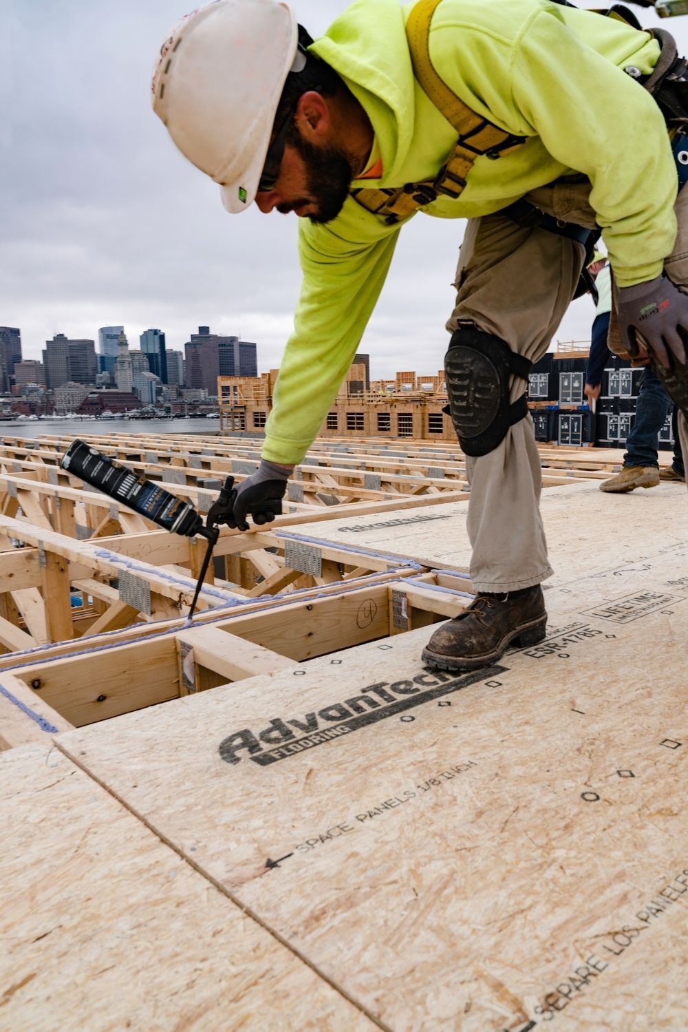 Construction worker using a caulk gun on wooden decking, city skyline in the background.