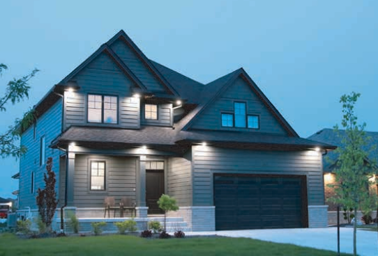 Two-story gray house with black roof and garage door, lit with outdoor lights at dusk.