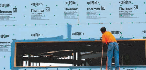 Construction worker in orange shirt and blue jeans leaning on a long tool, working on a building.