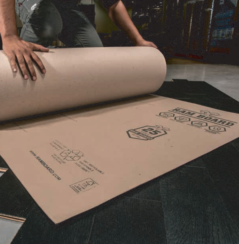 Person rolling out Ram Board protective paper on a dark floor; setting appears to be a construction site.