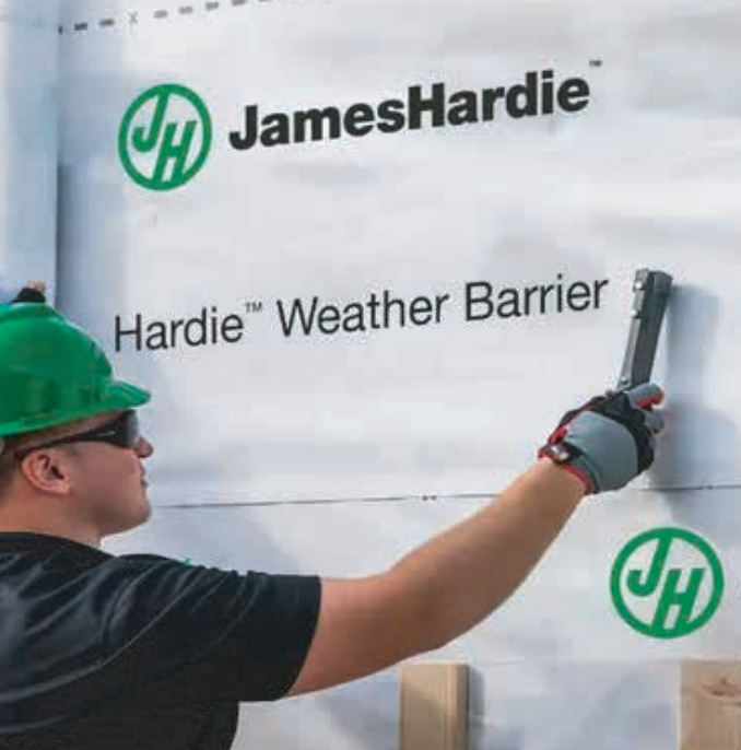 Construction worker installing James Hardie weather barrier. Green hard hat, gloves, black shirt. White barrier, green logo.