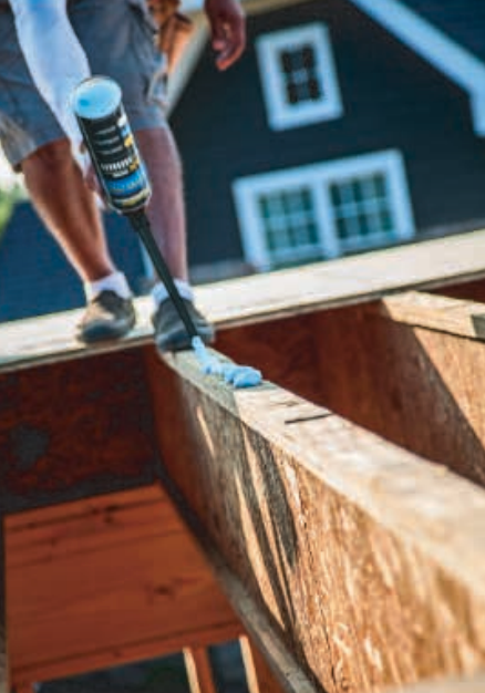 Person applying adhesive to a wooden beam on a construction site, house in the background.