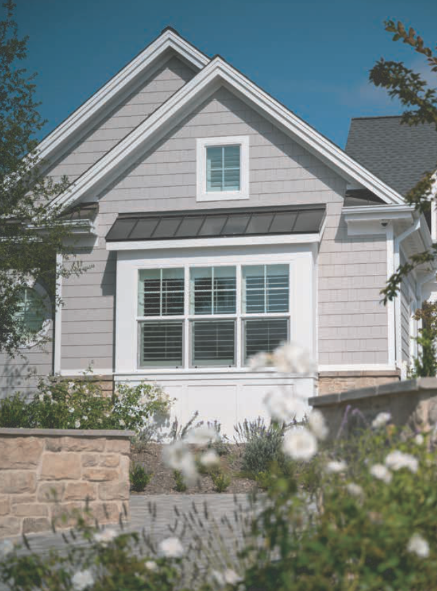 Gray house with white trim, window, and blue sky. Stone foundation, flowers in foreground.