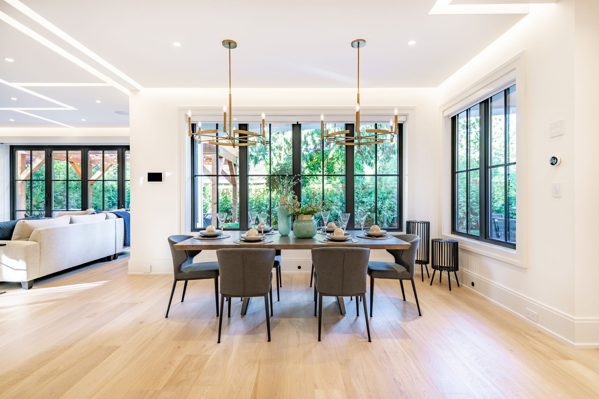 Elegant dining room with a rectangular table, six gray chairs, and large windows overlooking greenery.