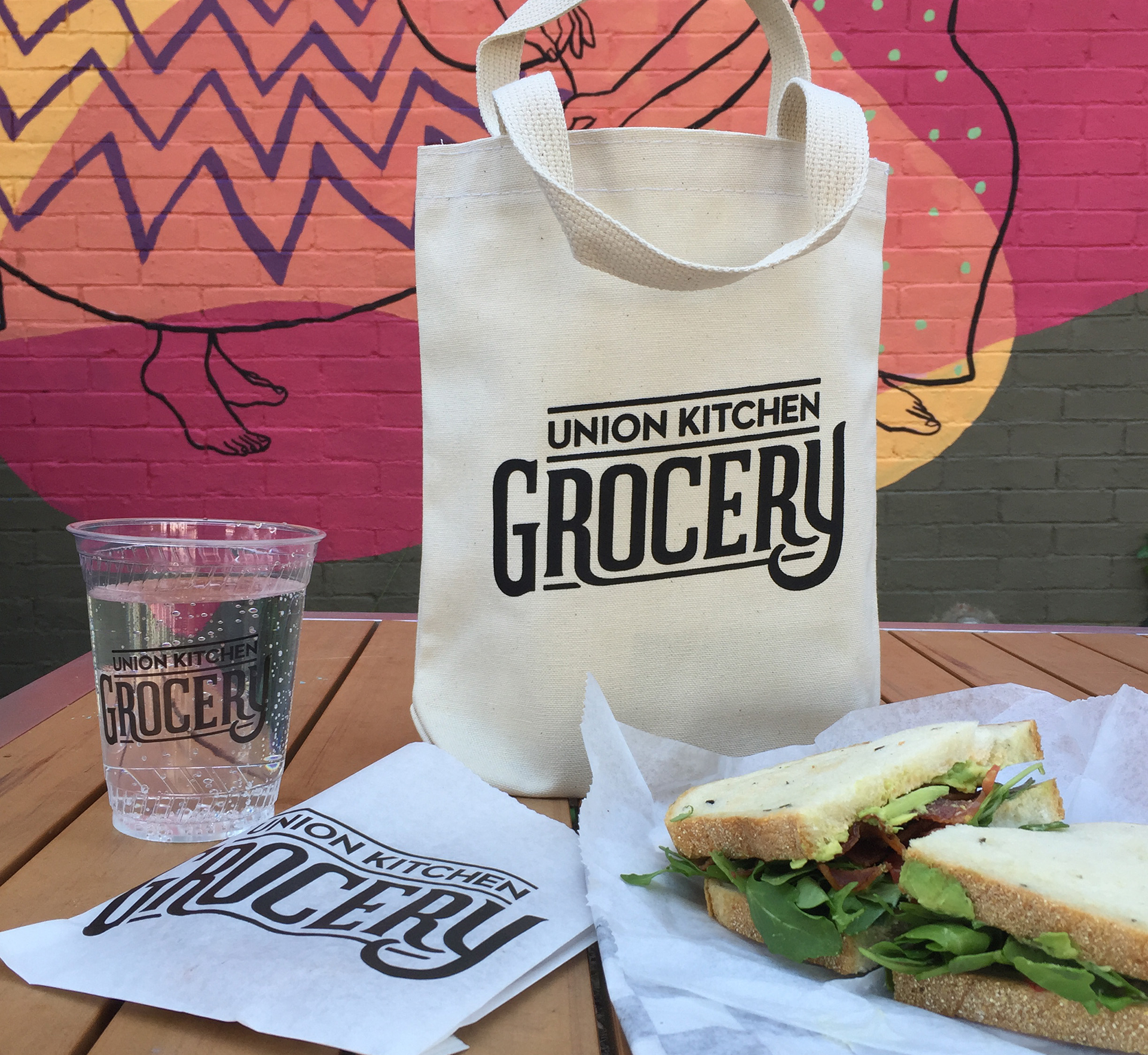 A grocery bag sits on a table next to a sandwich